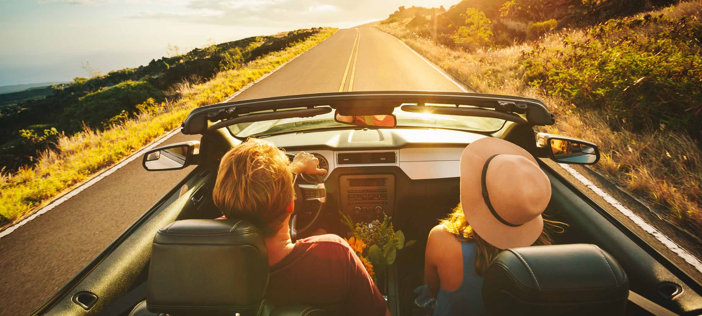 A back-view of a couple driving a black convertible on a winding coastal road at sunset, with golden light illuminating the hills and the road ahead - https://benderhatch.com/