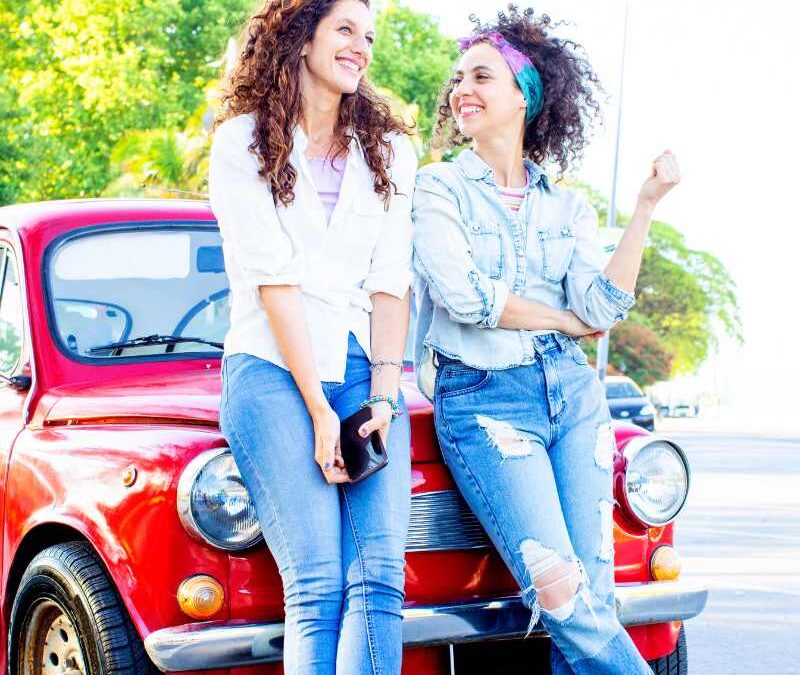 Two smiling women lean against a red vintage car